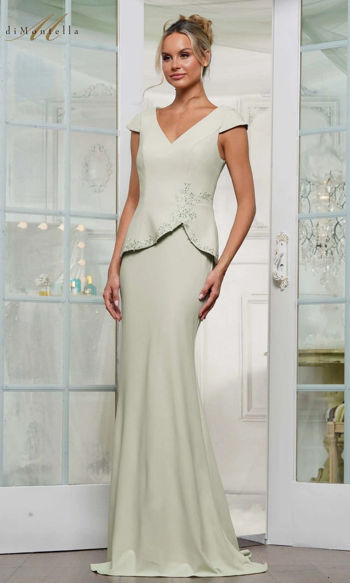 A woman stands indoors wearing the Rina Di Montella RD3239 long formal dress in light sage, featuring cap sleeves, a V-neck, subtle floral embroidery on the bodice, and a graceful mermaid skirt.